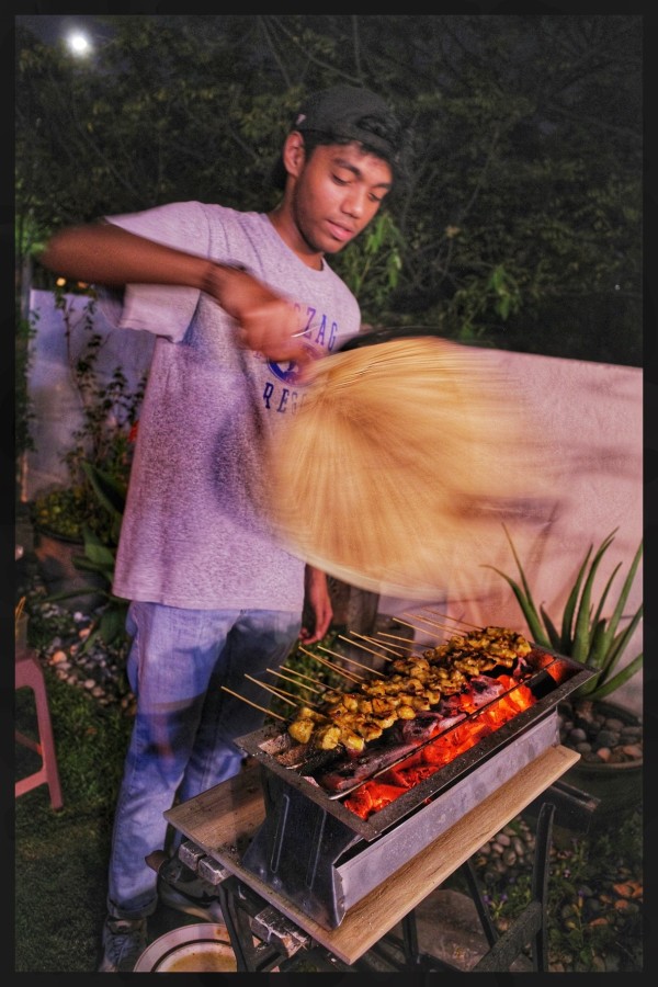 A man waves a fan above a rack of chicken skewers sitting on an open flame