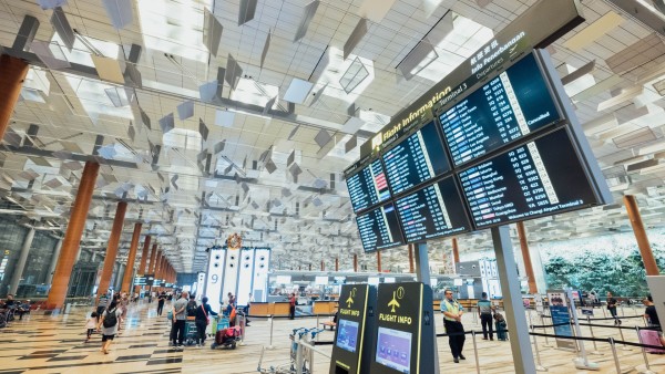 A large display board of flight details looms over an airport terminal